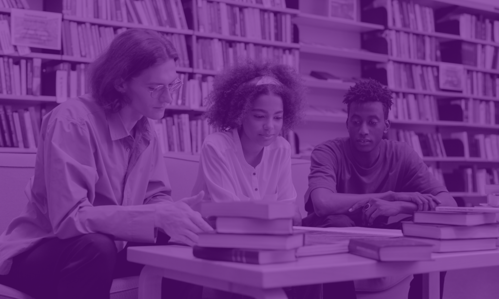 Three students studying in the library. Behind them is a wall covered by a bookcase. To the left is a man with brown shoulder length hair and glasses. In the center is a woman with an afro wearing a white shirt. To the right is a man with short curly hair wearing a dark t-shirt.