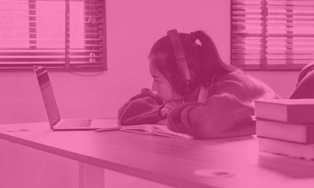 Young girl sitting at a desk revising on her laptop. She has her hair tied back and is wearing headphones. Her arms are crossed and she is leaning her chin on her arms. She has a confused expression.
