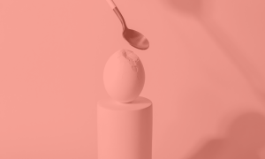 There is a boiled egg on a platform has been hit by a spoon (positioned above the egg). There is a dent and cracks in the right hand side of the shell. The image represents the phrase 'don't crack under pressure'